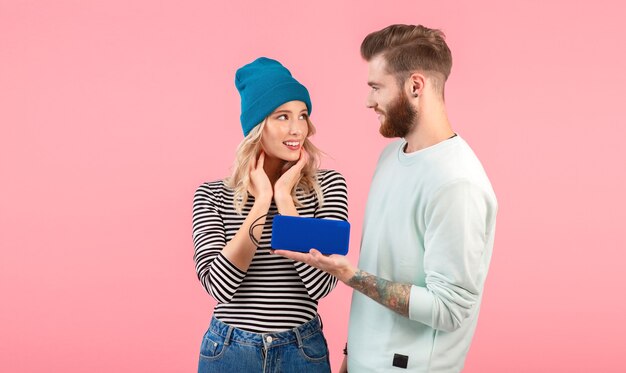 Young couple listening to music on wireless speaker wearing cool stylish outfit smiling posing on pink