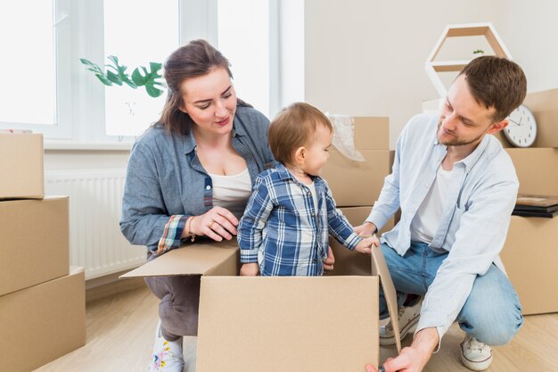 Young couple looking at their toddler son standing inside the cardboard box in the living room