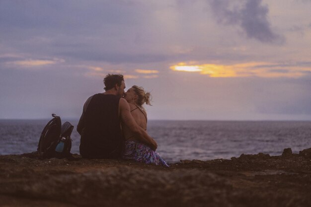 旅に恋をしている若いカップルが崖の端に座って夕日を眺めています。
