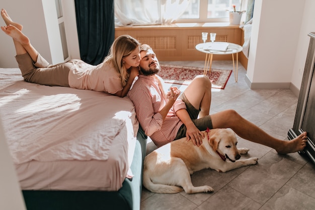 Young couple of lovers resting in bedroom. Girl in comfortable clothes lies on bed and looks at her boyfriend while he is stroking Labrador.