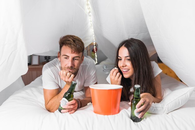 Молодая пара, лежа на кровати, наслаждаясь попкорн и алкогольные напитки во время просмотра телевизора