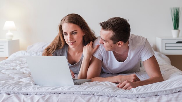 Молодая пара лежит на кровати с ноутбуком