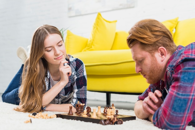 Young couple lying on carpet looking at chess board on the white carpet at home