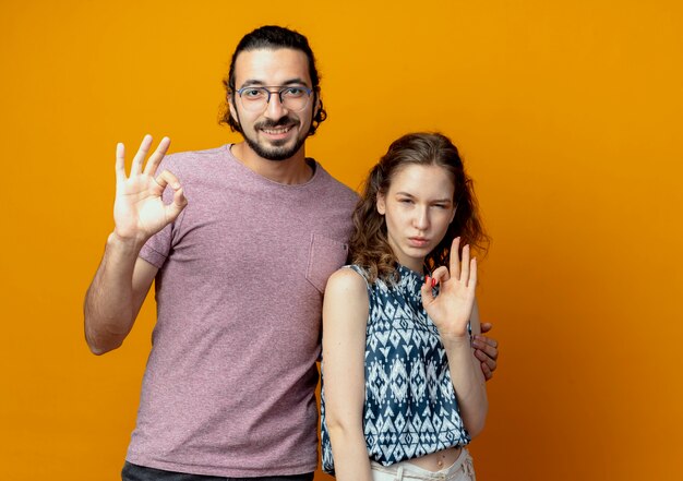 молодая пара делает знак ОК, улыбаясь с уверенным выражением лица, стоя над оранжевой стеной