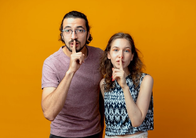young couple  making silence gesture with fingers on lips standing over orange wall
