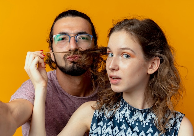 Бесплатное фото Молодая пара мужчина и женщина мужчина имитируют усы с волосами своей подруги над оранжевой стеной