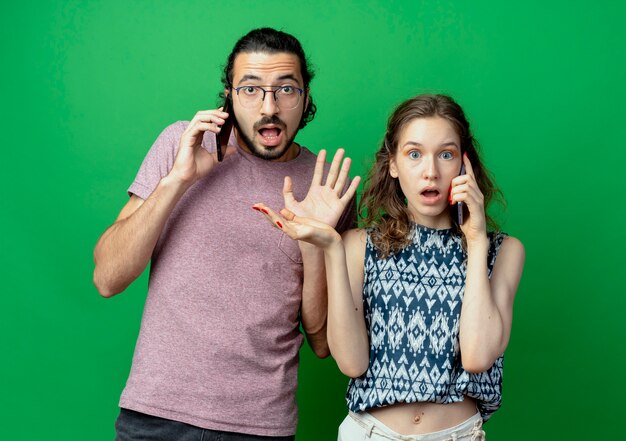 Молодая пара мужчина и женщина были шокированы и разочарованы во время разговора по мобильным телефонам, стоя на зеленом фоне