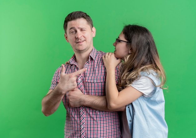 녹색 벽 위에 서있는 그녀의 여자 친구에게 손가락으로 가리키는 캐주얼 옷 자신감 남자 젊은 부부 남자와 여자