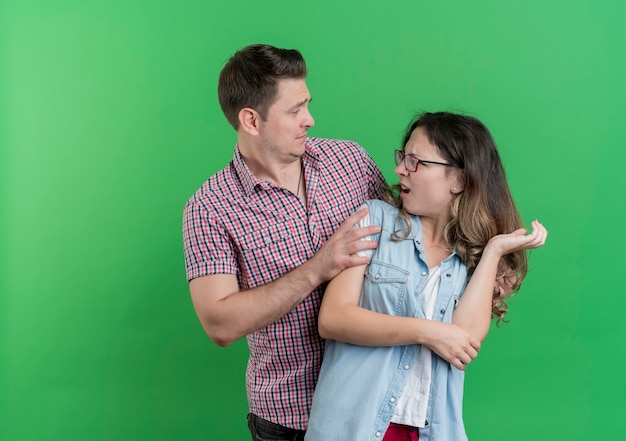 カジュアルな服を着た若いカップルの男性と女性は、緑の壁の上に立っている彼の怒っているガールフレンドに許しを求めて混乱した男