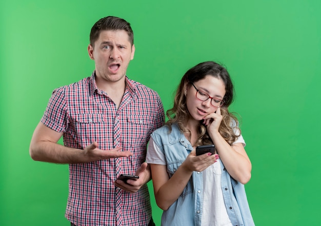 カジュアルな服を着た若いカップルの男性と女性は、緑の壁の上に立っている携帯電話で話している彼の忙しいgirlfrinedを腕で指して混乱した男