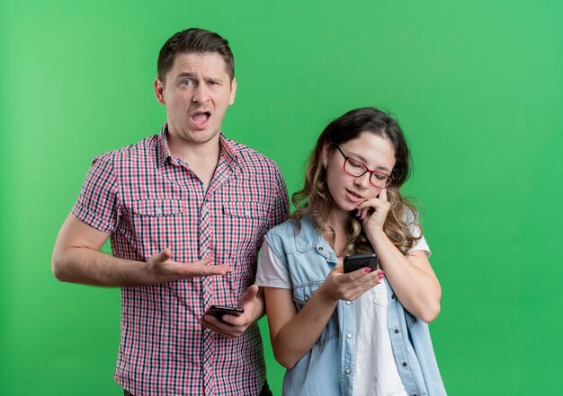 젊은 커플 남자와 캐주얼 옷에 여자는 녹색 벽 위에 서있는 휴대 전화에 말하는 그의 바쁜 girlfrined에서 팔을 가리키는 남자를 혼란