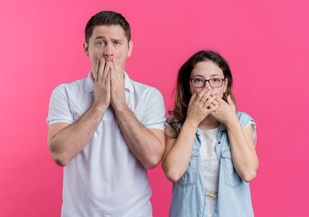 Young couple man and woman in casual clothes covering mouth with hands being shocked over pink