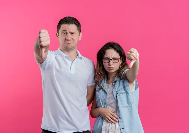 Молодая пара мужчина и женщина в повседневной одежде недовольны, показывая большие пальцы вниз, стоя над розовой стеной