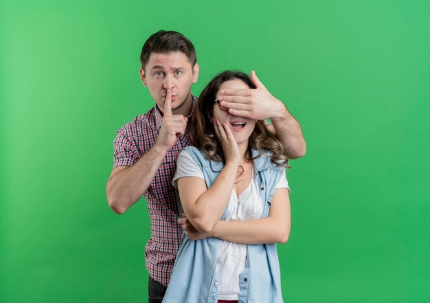 Young couple man and woman in casual clothes happy man covering his girlfriends eyes making a surprise with finger on lips over green
