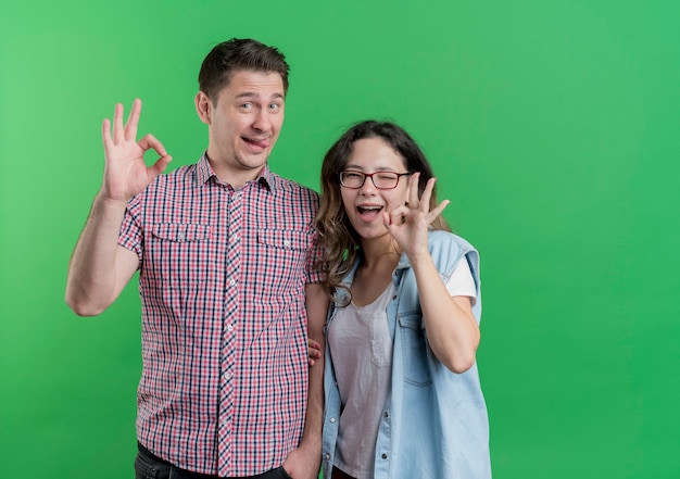 Young couple man and woman in casual clothes happy and positive showing ok signs having fun together standing over green wall