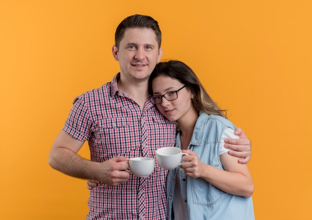 オレンジ色の壁の上に幸せで前向きに立っているコーヒーカップを保持しているカジュアルな服を着た若いカップルの男性と女性