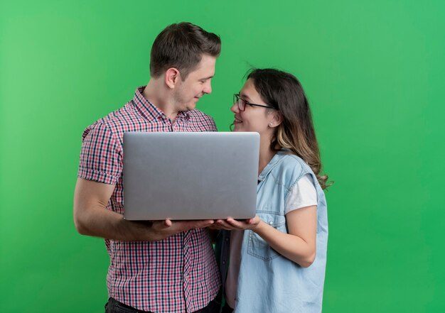 Молодая пара мужчина и женщина в повседневной одежде, держа ноутбук, глядя друг на друга, улыбаясь, стоя над зеленой стеной