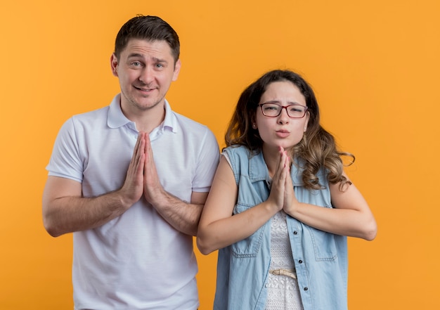 Young couple man and woman in casual clothes holding palms together making wish with hope expression standing over orange wall
