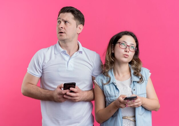 Young couple man and woman in casual clothes holding smartphones looking asides with pensive expression standing over pink wall