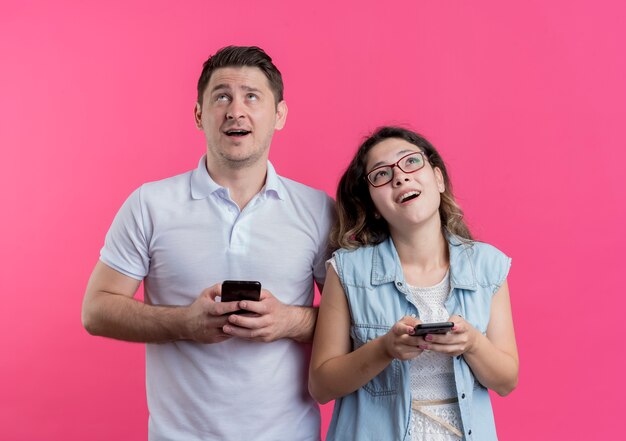 Young couple man and woman in casual clothes holding smartphones looking up with dreamy look standing over pink wall