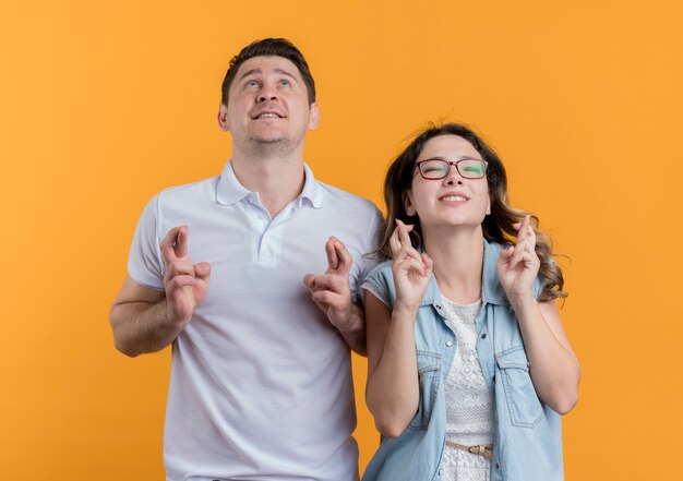 Young couple man and woman in casual clothes looking up making desirable wish crossing fingers over orange