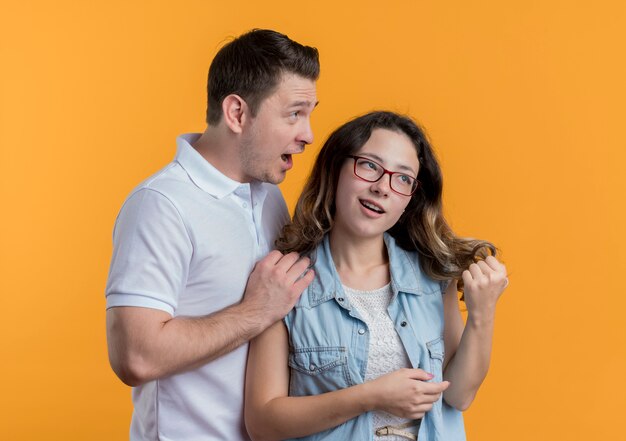 Young couple man and woman in casual clothes man talking to his girlfriend looking surprised over orange