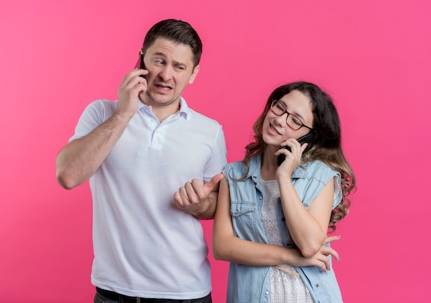 ピンクの上に彼女のガールフレンドを指して混乱しているように見える携帯電話で話しているカジュアルな服を着た若いカップルの男性と女性