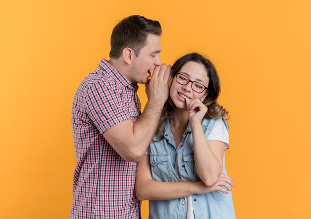 Young couple man and woman in casual clothes man whispering a secret to his smiling girlfriend standing over orange wall