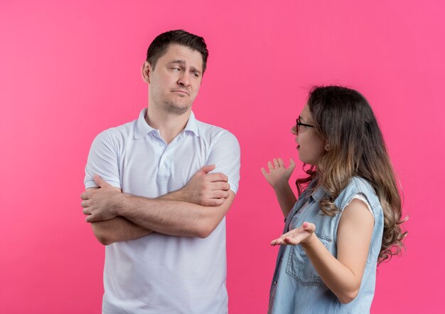 Young couple man and woman in casual clothes man with crossed hands on chest while his girlfriend arguing with him standing over pink wall