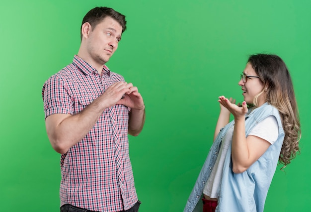 Free photo young couple man and woman in casual clothes sad man showing heart gesture to her displeased girlfriend standing over green wall