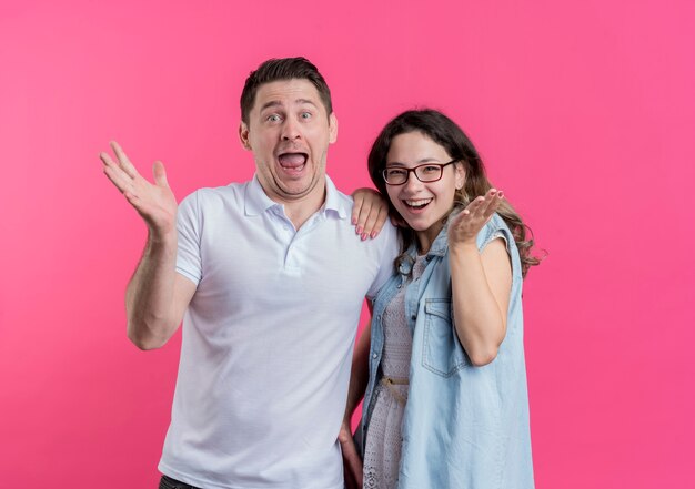 Young couple man and woman in casual clothes  smiling cheerfully happy and surprised standing over pink wall