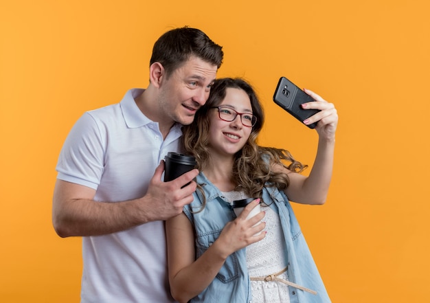 Coppia giovane uomo e donna in abiti casual che stanno insieme facendo selfie felice nell'amore divertendosi sopra la parete arancione