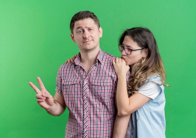 Young couple man and woman in casual clothes standing together hapy man showing v-sign over green wall