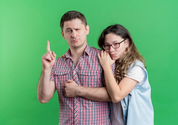 Молодая пара мужчина и женщина в повседневной одежде, стоящие вместе уверенным в себе мужчиной, показывающим указательный палец над зеленой стеной