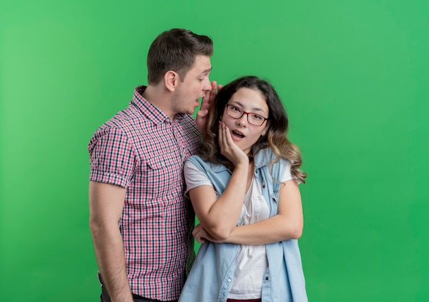 Young couple man and woman in casual clothes surprised man whispering a secret to his amazed girlfriend standing over green wall