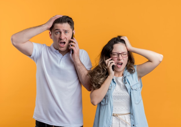 Young couple man and woman in casual clothes talking on mobile phones looking confused and very anxious touching their heads standing over orange wall