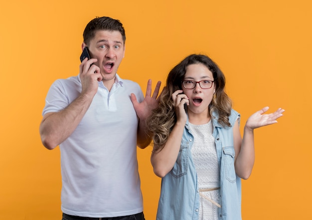 Молодая пара мужчина и женщина в повседневной одежде разговаривают по мобильным телефонам, выглядят смущенными и обеспокоенными, стоя у оранжевой стены