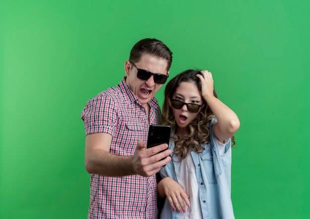 緑の壁の上に立って驚いて混乱しているスマートフォンの画面を見て黒い眼鏡をかけているカジュアルな服を着た若いカップルの男性と女性