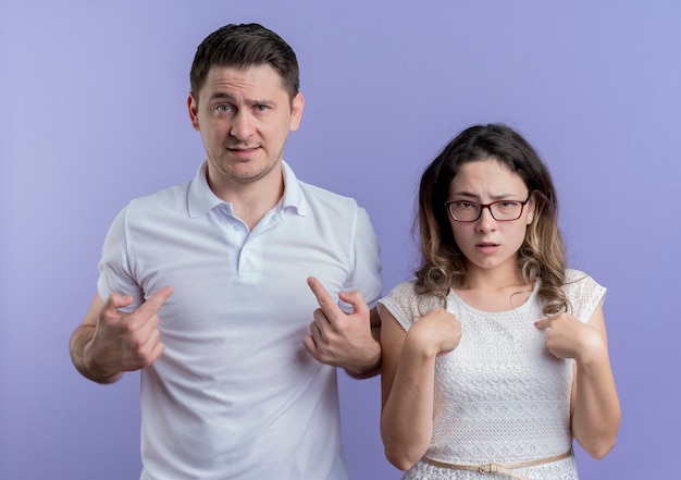 Young couple man and woman confused pointign with fingers at themselves over blue