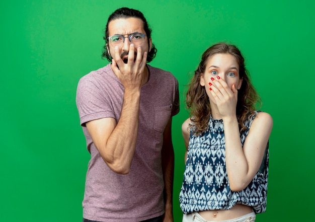 young couple man and woman,  covering mouthes with hands being shocked standing over green wall