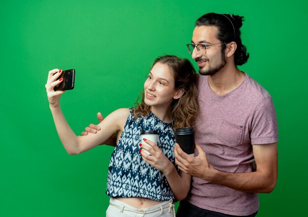 young couple man and woman happy in love, happy woman taking picture of them using smartphone standing over green wall