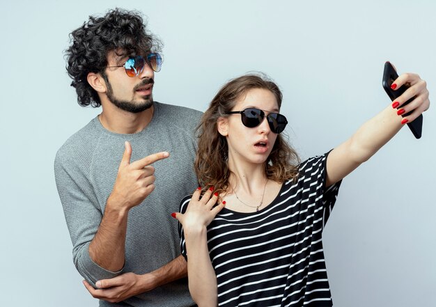 young couple man and woman happy in love, happy woman taking picture of them using smartphone standing over white wall