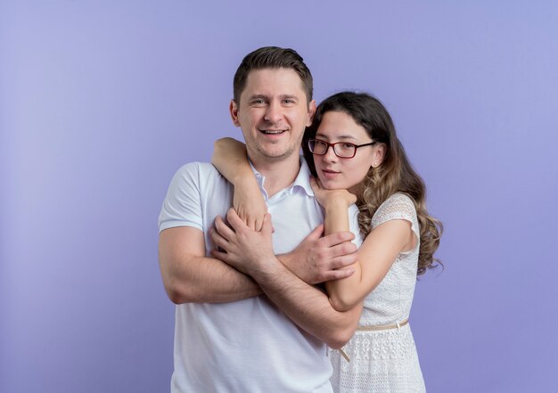Young couple man and woman happy in love huging looking at camera smiling standing over blue wall