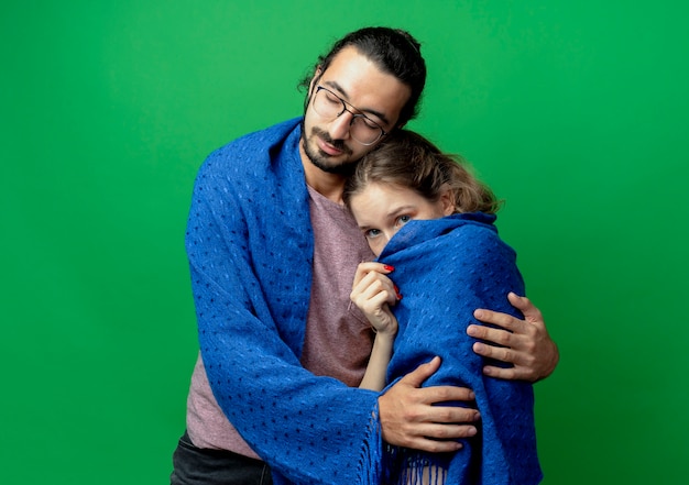 Молодая пара мужчина и женщина, счастливый мужчина обнимает свою любимую подругу, завернув ее в теплое одеяло, стоящее на зеленом фоне