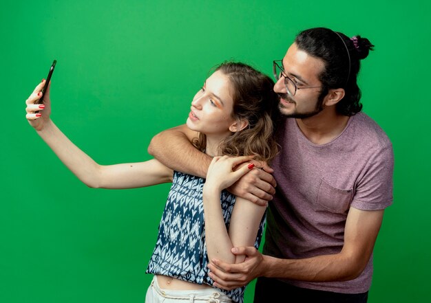 若いカップルの男性と女性、緑の背景の上に立っている彼女のスマートフォンを使用してそれらの写真を撮る幸せな女性