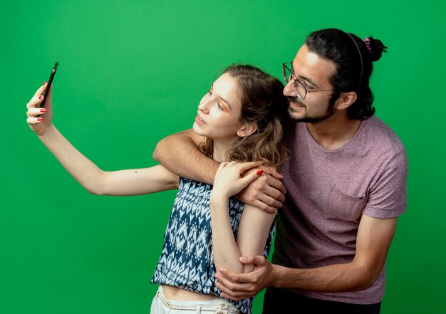 Young couple man and woman, happy woman taking picture of them using her smartphone standing over green background