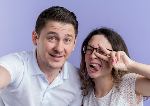 Young couple man and woman  having fun together showing v-sign standing over blue wall