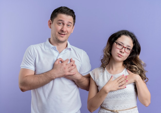 Young couple man and woman holding hands on chest feeling thankful happy and positive standing over blue wall