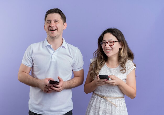 スマートフォンを持っている若いカップルの男性と女性が青い壁の上に元気に一緒に立って幸せで前向きな笑顔