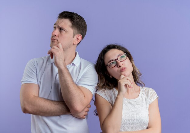 Young couple man and woman looking asides with pensive expression on face standing over blue wall